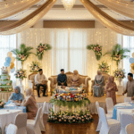 A detailed wide-angle photograph capturing a formal Islamic baby shower, or Aqiqah (cradle ceremony) celebration hall. The scene is set in an elegantly decorated hotel banquet room, featuring draped cream and gold fabrics with fairy lights on the ceiling and a central crystal chandelier. An elevated stage with a lit backdrop displays ornate sofas where four elders are seated. A central decorative cradle with a sleeping baby is nestled within a large ring of flowers on a pedestal. Around the tables in the foreground, family members and guests, dressed in traditional and modest attire (including hijabs), are gathered. A large multi-tiered pasture-themed Aqiqah cake with balloons is on the left, and a wicker goat figurine is on the right. Warm ambient lighting fills the room, and all details, including table settings with blue runners, are clear.
