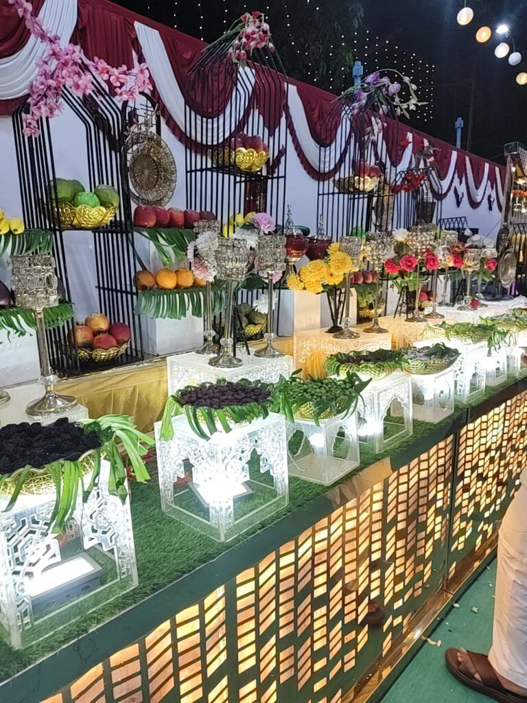 An elaborate outdoor catering buffet line featuring glowing, backlit white display stands filled with fresh greens, topped by tall black metal tier structures displaying fresh fruits, decorative gold platters, and floral arrangements against a red and white draped background.