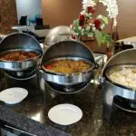 Four stainless steel roll-top chafing dishes with open lids arranged in a row on a dark marble countertop, containing white rice, fried noodles, and assorted meat and vegetable entrees next to empty white side plates.