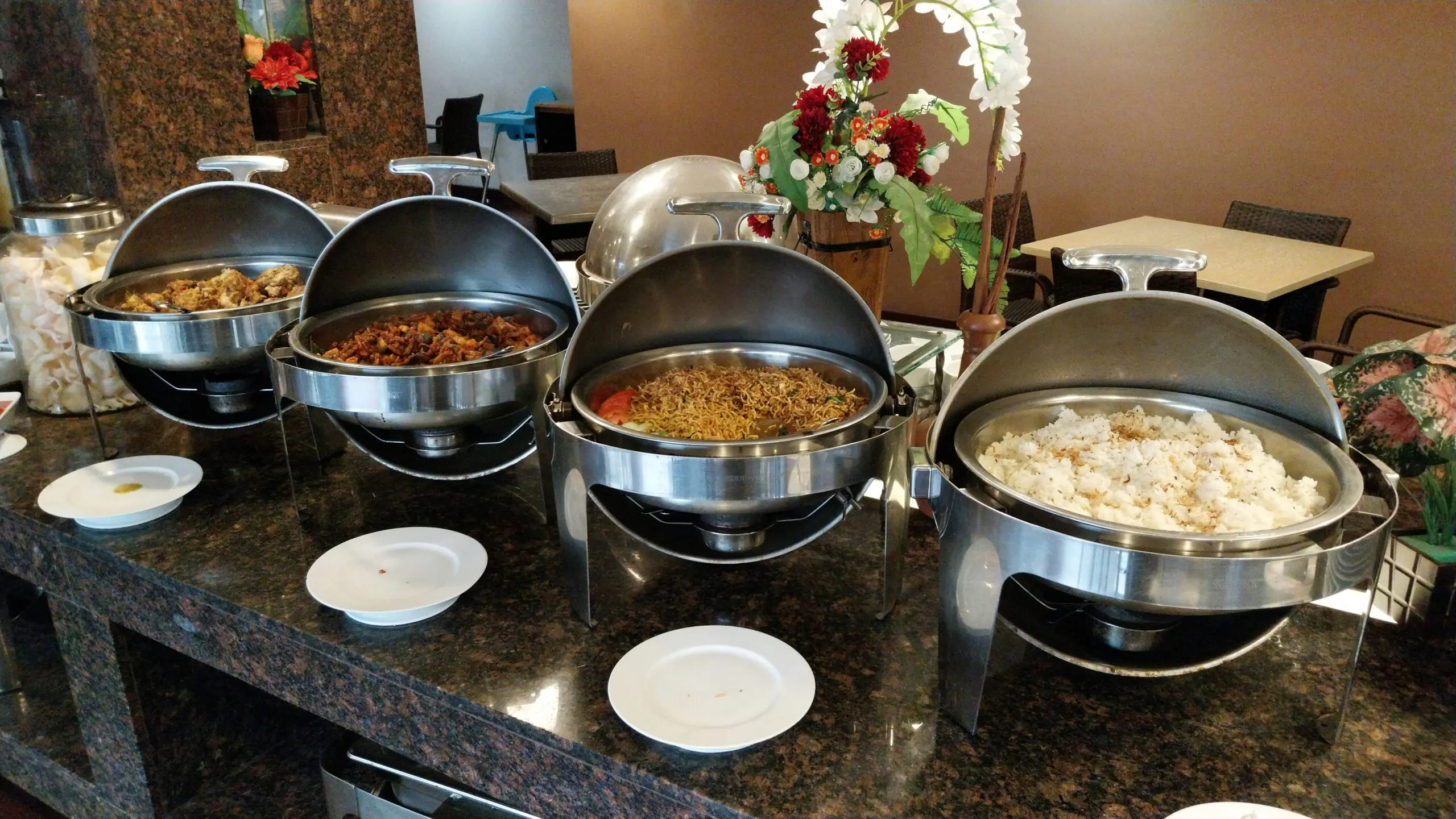 Four stainless steel roll-top chafing dishes with open lids arranged in a row on a dark marble countertop, containing white rice, fried noodles, and assorted meat and vegetable entrees next to empty white side plates.