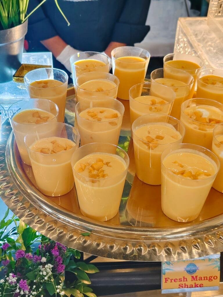 A high-angle shot showing multiple clear plastic glasses filled with creamy, yellow Fresh Mango Shake arranged neatly on a large, reflective silver platter at a luxury catering event buffet. Each mini dessert cup is topped with small pieces of diced fresh mango. In the bottom right corner, a small printed sign reads 'Fresh Mango Shake' with a blue 'Food Forest Catering' logo visible on it.