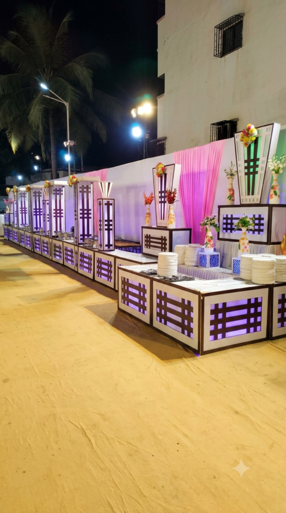 A vertical 9:16 portrait photograph capturing a meticulously arranged and polished outdoor catering display at night. The long buffet line, featuring white panels with brown lattice-work and internally lit in soft purple and white, extends from the foreground to the background. Vases with fresh flowers are strategically placed along the line, and white plates are neatly stacked. In the background, the large, backlit V-shaped structures with floral arrangements are visible. The pink drapery and background structures are clean and clear. The sandy ground is smooth and empty, without any chairs or clutter. The overhead lighting is warm and balanced.