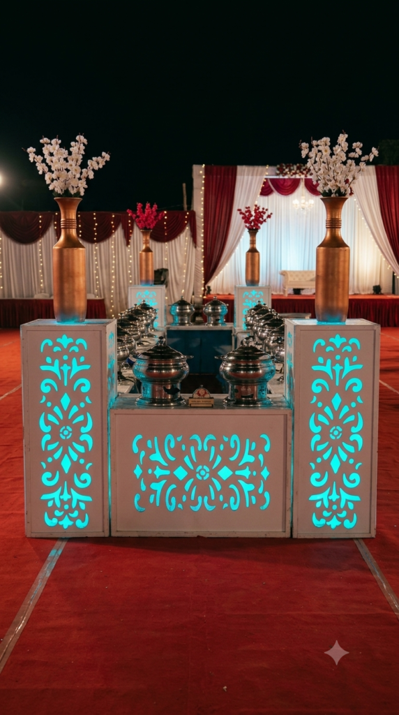 A professional evening catering buffet setup at an outdoor event venue. In the foreground, white counter panels feature intricate laser-cut floral patterns glowing with brilliant teal backlighting. Tall, metallic bronze vases holding branches of white blossoms stand symmetrically atop the illuminated pillars. Behind them, polished stainless steel chafing dishes are neatly arranged on a red carpet. The background features a draped stage with white and crimson curtains under a dark night sky.
