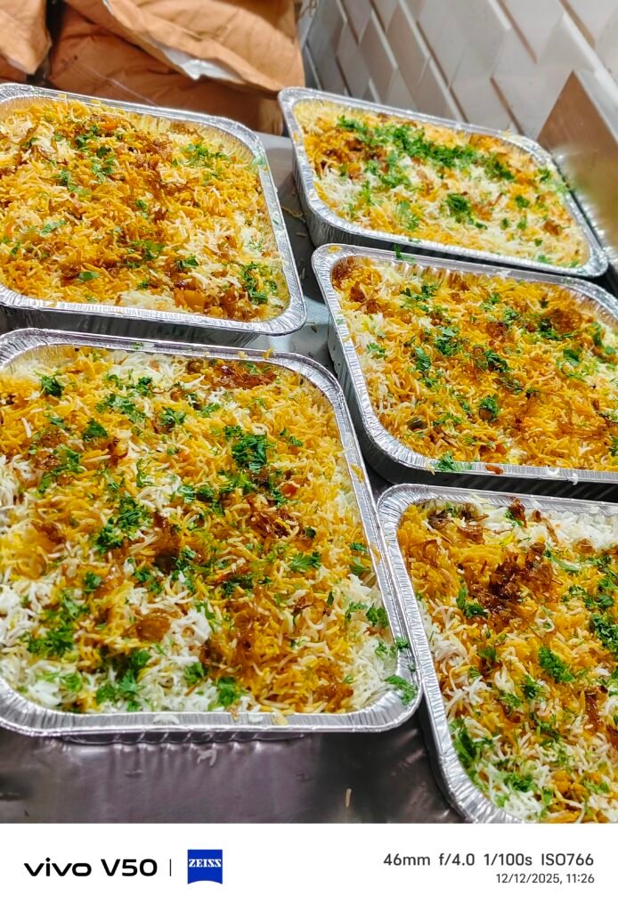 biryani catering mumbai A high-resolution, top-down shot of a large, rectangular aluminum foil container filled with a vibrant vegetable biryani. The dish displays distinct layers of fluffy white and bright yellow-orange basmati rice mixed with peas and carrots. It is heavily garnished across the surface with golden-brown fried onions and scattered fresh green coriander leaves.